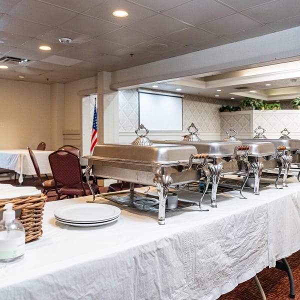 A buffet line with several silver chafing dishes, plates, and hand sanitizer on a white-covered table.