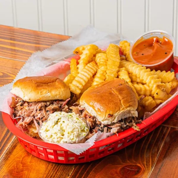 Two pulled pork sandwiches, crinkle-cut fries, coleslaw, and BBQ sauce in a red basket.
